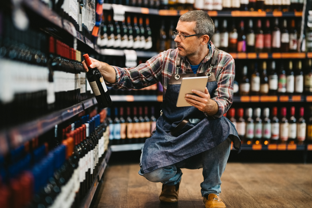 Mature salesman with digital tablet taking stock of liquor in wine section at grocery store
